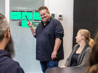 René zeigt mit einem Stift auf Klebezetteln. Menschen sitzen im Vordergrund und haben Spaß.