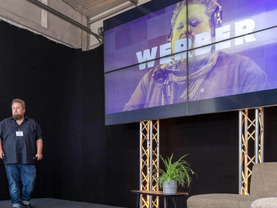 René auf einer Bühne. TV im Hintergrund zeigt einen Schriftzug: Werber.