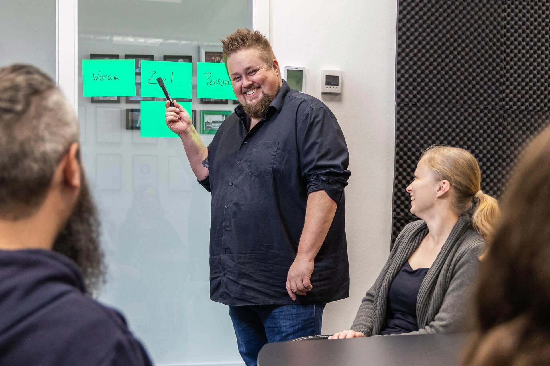 René zeigt mit einem Stift auf Klebezetteln. Menschen sitzen im Vordergrund und haben Spaß.
