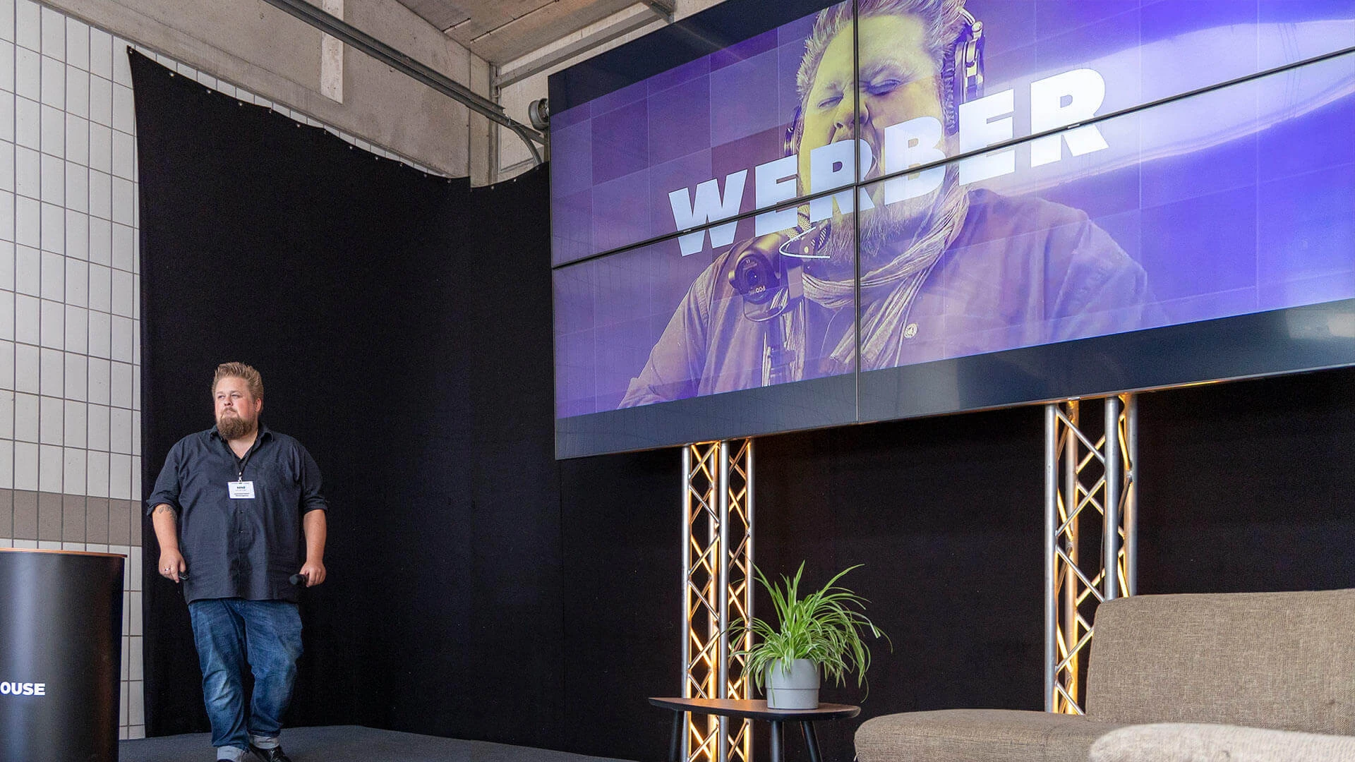 René auf einer Bühne. TV im Hintergrund zeigt einen Schriftzug: Werber.