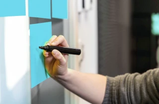 Klebezettel an einer Wand. Eine Hand mit einem Stift.